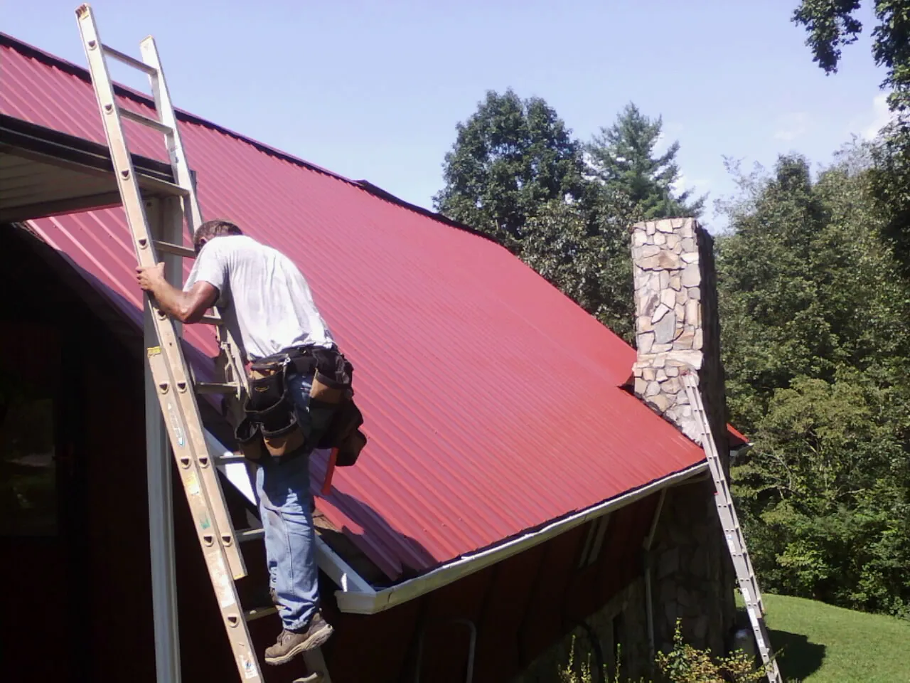 Roofer on a ladder beside a red metal roof and stone chimney — Soffit & Fascia Repair by Roofing Brothers in Big Prairie